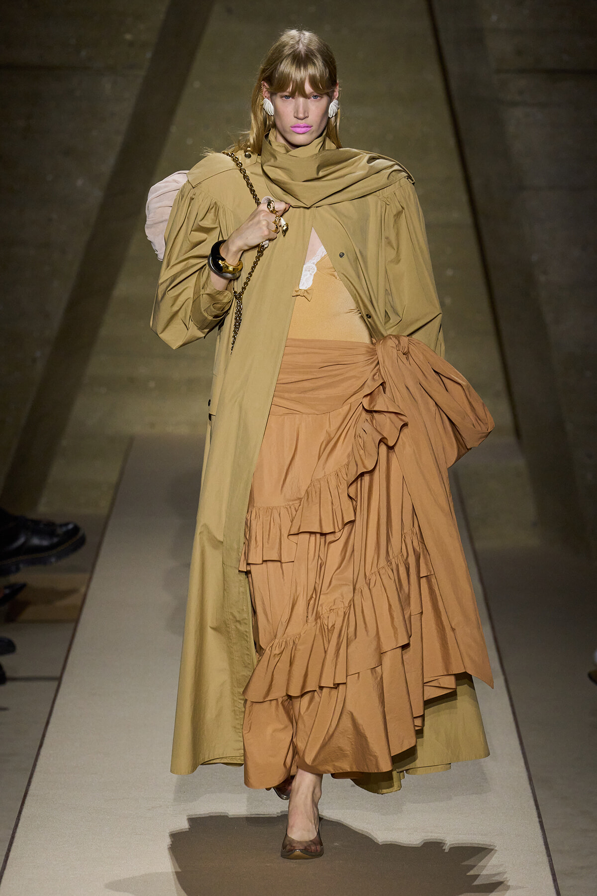 Model walking a runway in a tan trench over a layered camel dress with ruffles and an oversized bow detail at the waist, wearing nude heels and bold pink lipstick.