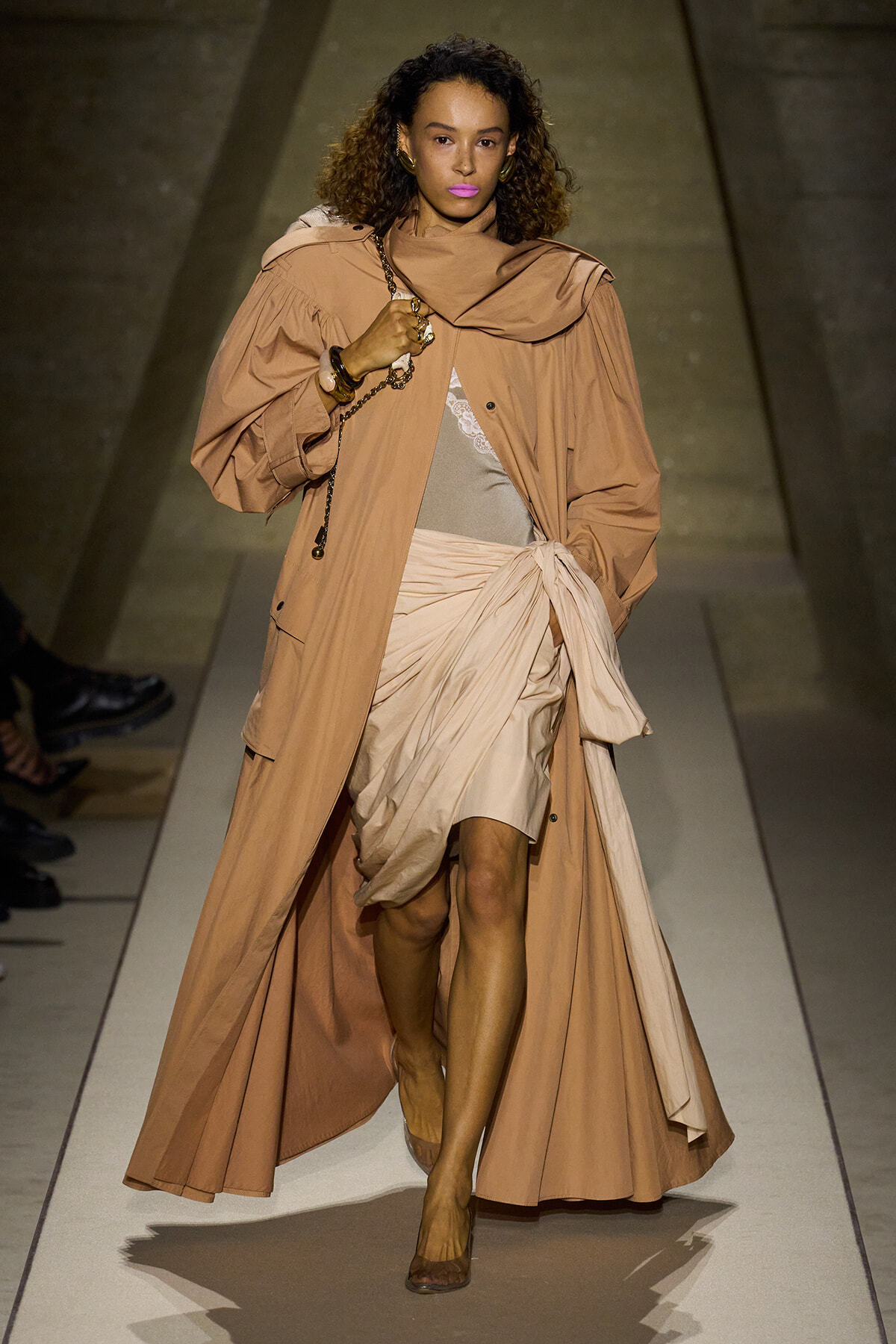 Model walking a fashion runway wearing a tan oversized trench and wrapped beige dress with matching scarf-like detail.