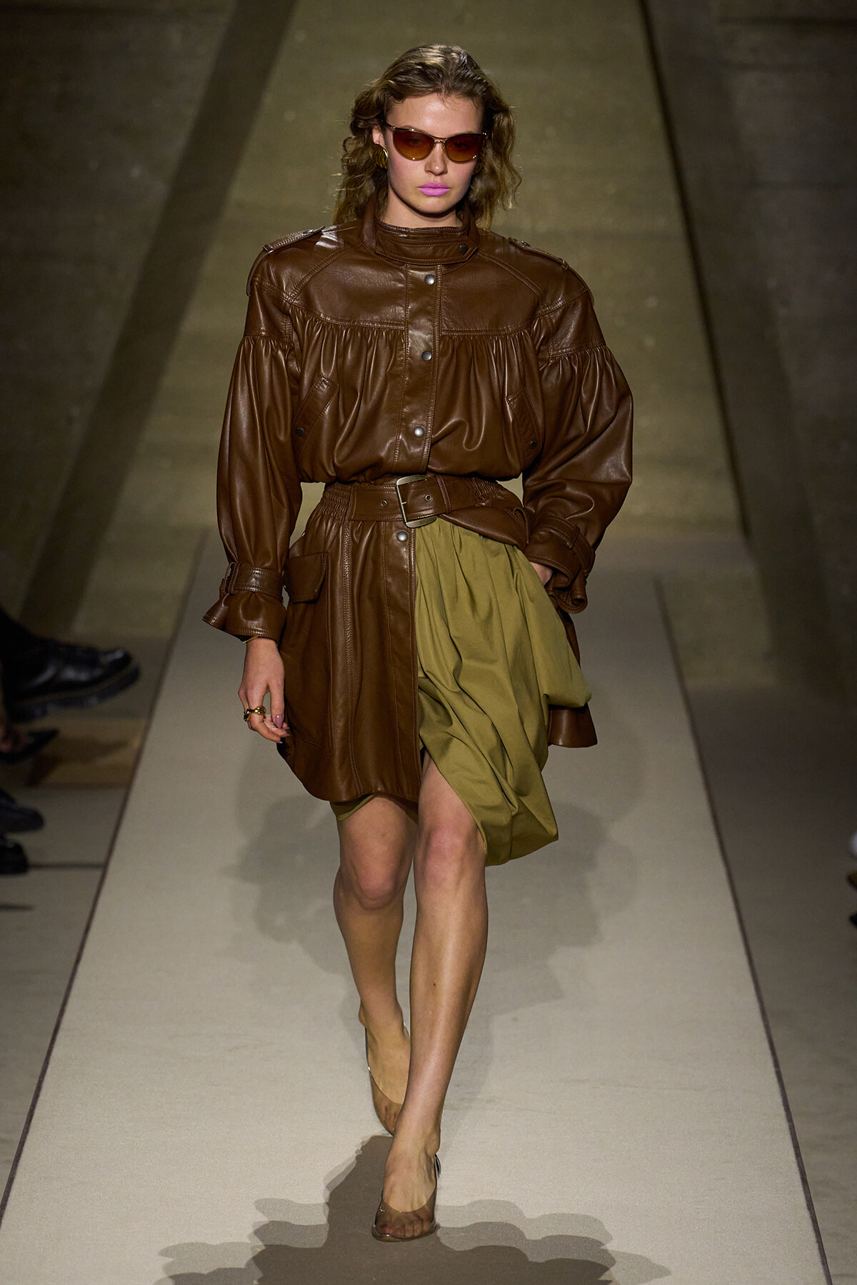 Female model walking a fashion runway in a brown leather oversized jacket cinched at the waist with a belt, olive draped skirt, and brown heel shoes, wearing tinted sunglasses.