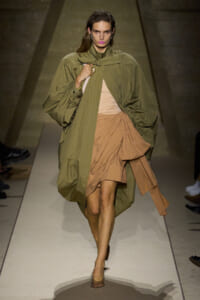 Model walks a runway in an oversized olive trench layered over a tan draped dress with a knot at the waist; she carries a light bag and wears minimal accessories.