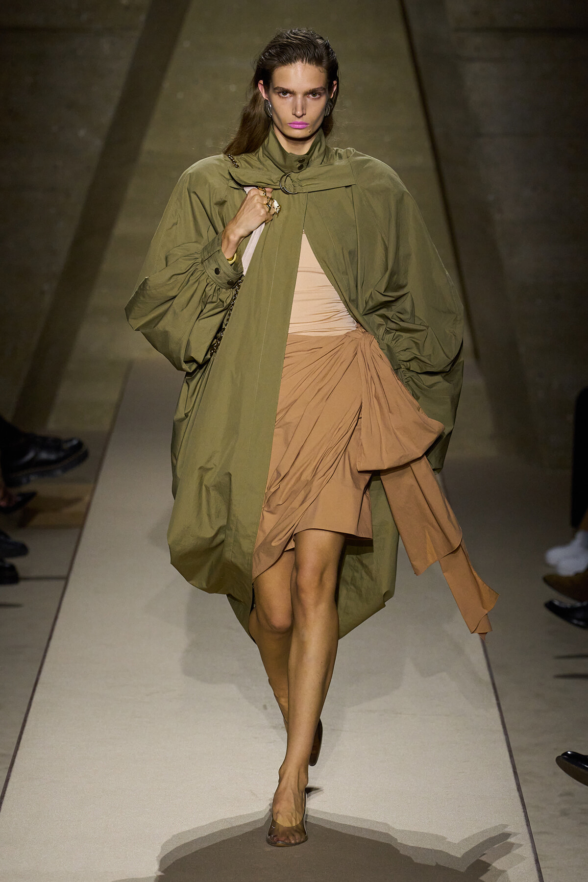 Model walks a runway in an oversized olive trench layered over a tan draped dress with a knot at the waist; she carries a light bag and wears minimal accessories.
