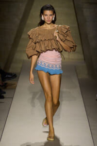 Model walks the runway in a tan oversized ruffled blouse and pastel pink and blue lace-trim shorts, carrying a chain-strap bag.