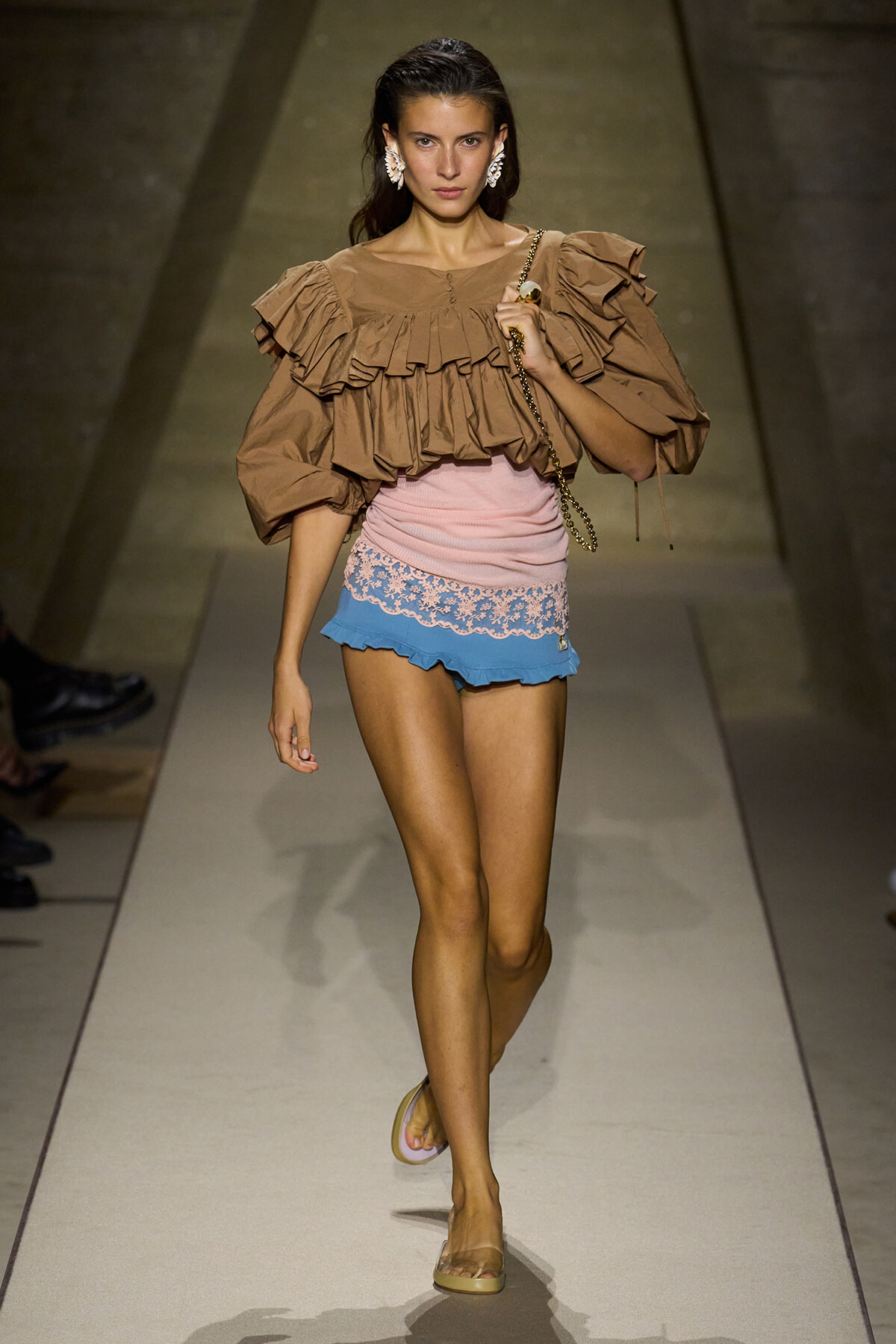 Model walks the runway in a tan oversized ruffled blouse and pastel pink and blue lace-trim shorts, carrying a chain-strap bag.