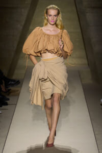 Model walking the runway in a camel-beige off-the-shoulder puff-sleeve crop top and matching draped wrap skirt on a fashion catwalk.