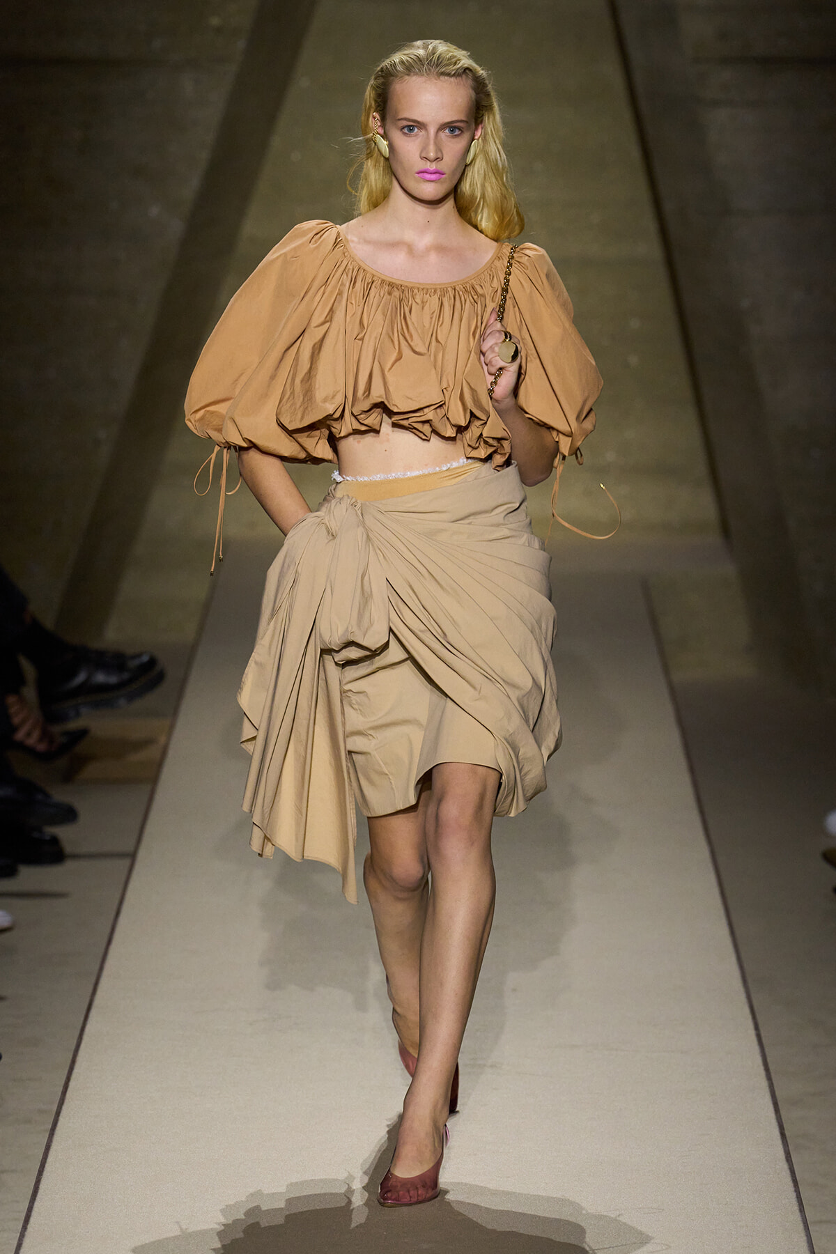 Model walking the runway in a camel-beige off-the-shoulder puff-sleeve crop top and matching draped wrap skirt on a fashion catwalk.