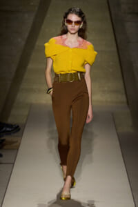 Model on a runway wearing a yellow off-shoulder knit top with pink lace collar, brown high-waisted pants, and a wide gold belt with yellow sandals.