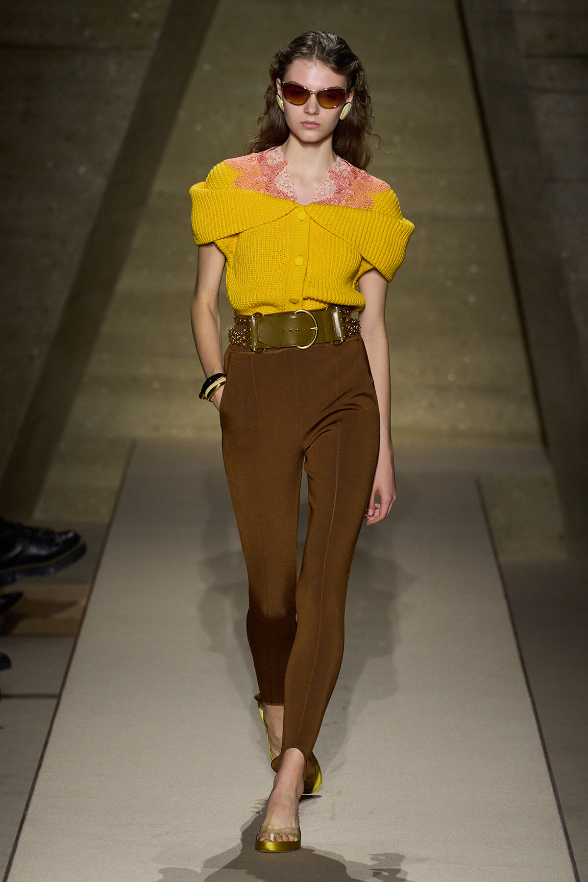 Model on a runway wearing a yellow off-shoulder knit top with pink lace collar, brown high-waisted pants, and a wide gold belt with yellow sandals.