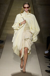 Model walking a runway in a pale cream oversized coat and draped dress, accessorized with a chain bag and sunglasses.