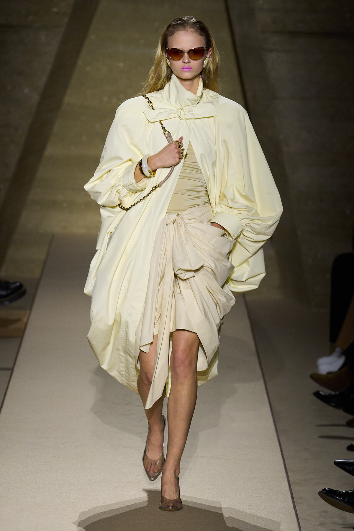 Model walking a runway in a pale cream oversized coat and draped dress, accessorized with a chain bag and sunglasses.