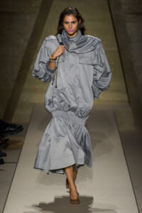 Model walks the runway in an oversized light-gray coat dress with a cinched waist and large puff sleeves.