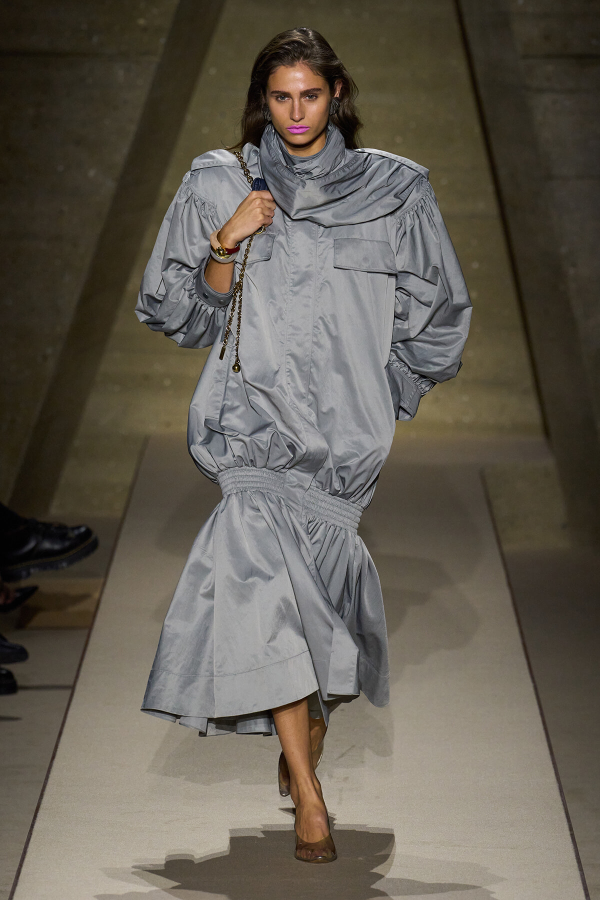 Model walks the runway in an oversized light-gray coat dress with a cinched waist and large puff sleeves.