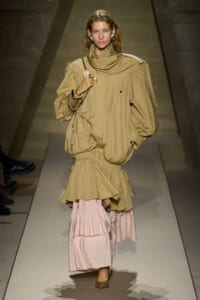 Model on a runway wearing a tan oversized jacket with a pink tiered skirt and a chain bag accessory.