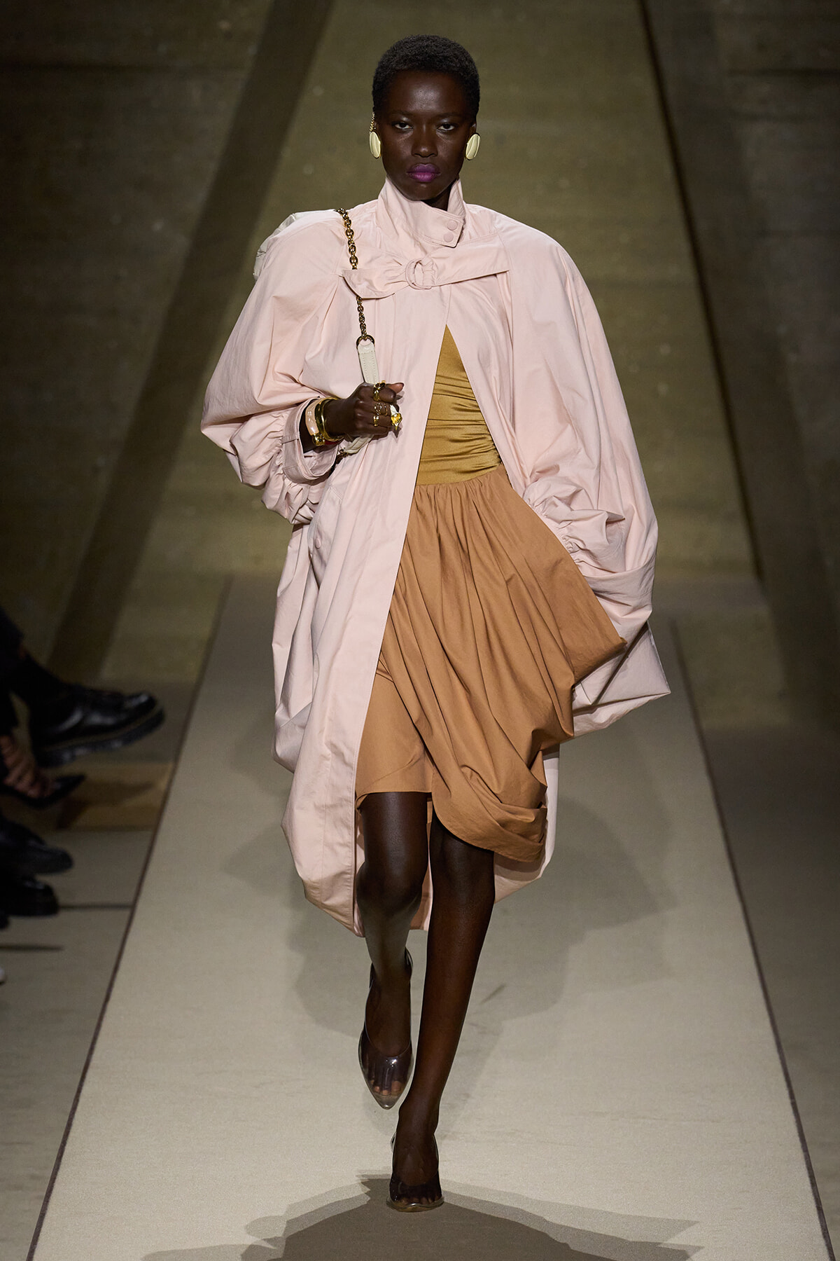 Model walking a runway in a pale pink oversized cape over a tan gathered dress with gold jewelry and large white earrings.