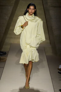 Model walks a runway in a pale yellow oversized outfit with a draped scarf and matching shoes, carrying a chain-strap bag on her shoulder.