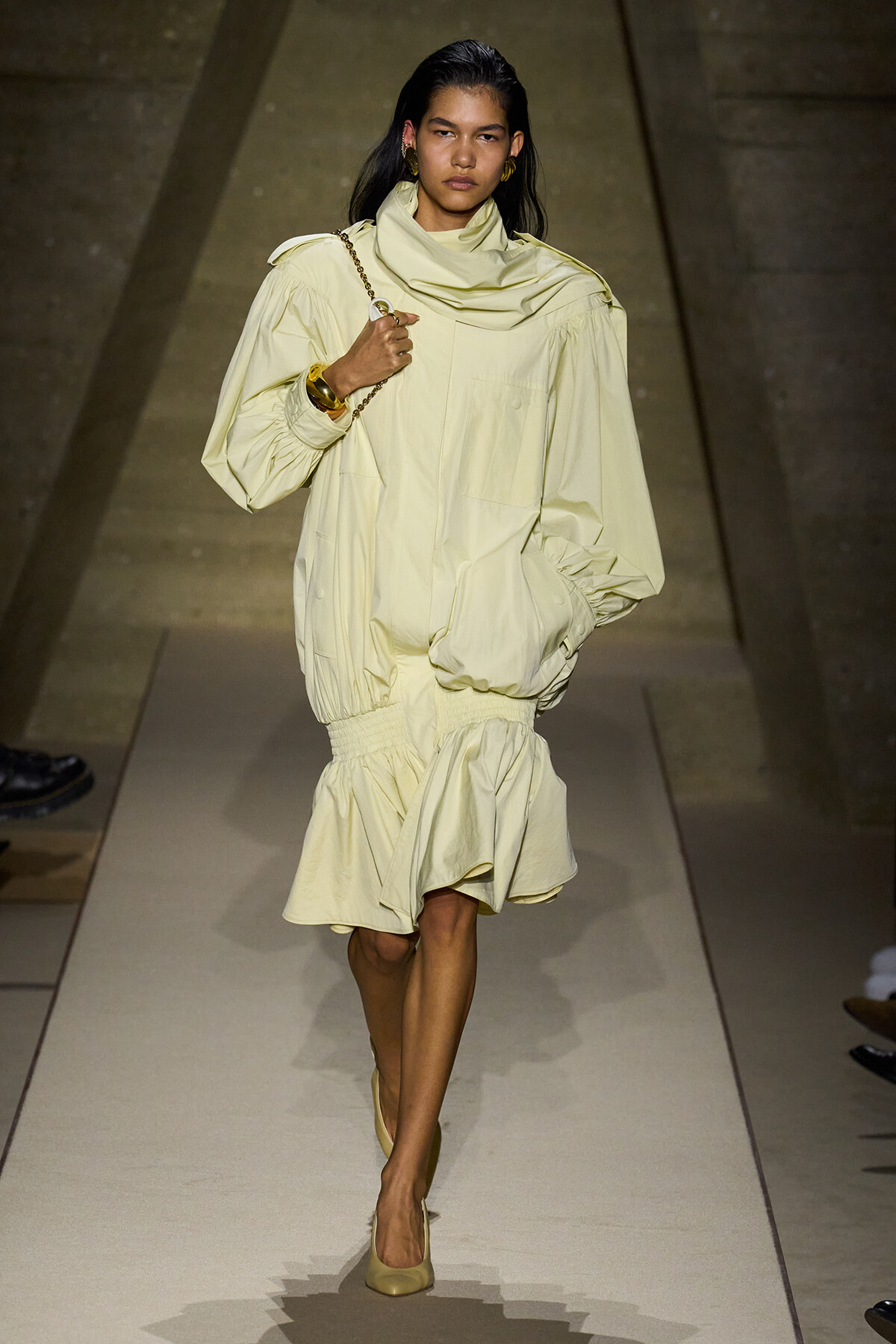 Model walks a runway in a pale yellow oversized outfit with a draped scarf and matching shoes, carrying a chain-strap bag on her shoulder.