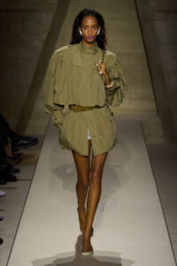 Model walks the runway in an olive-green, oversized cape-style top with a matching belt and shorts, carrying a chain bag.