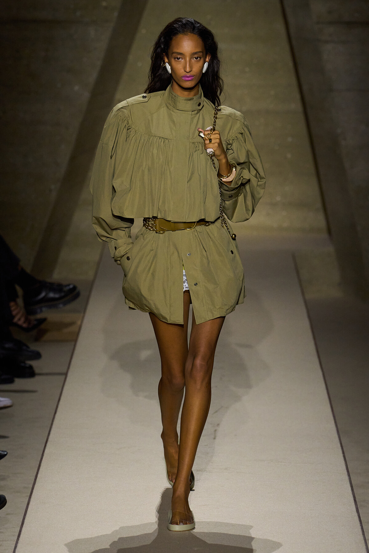 Model walks the runway in an olive-green, oversized cape-style top with a matching belt and shorts, carrying a chain bag.