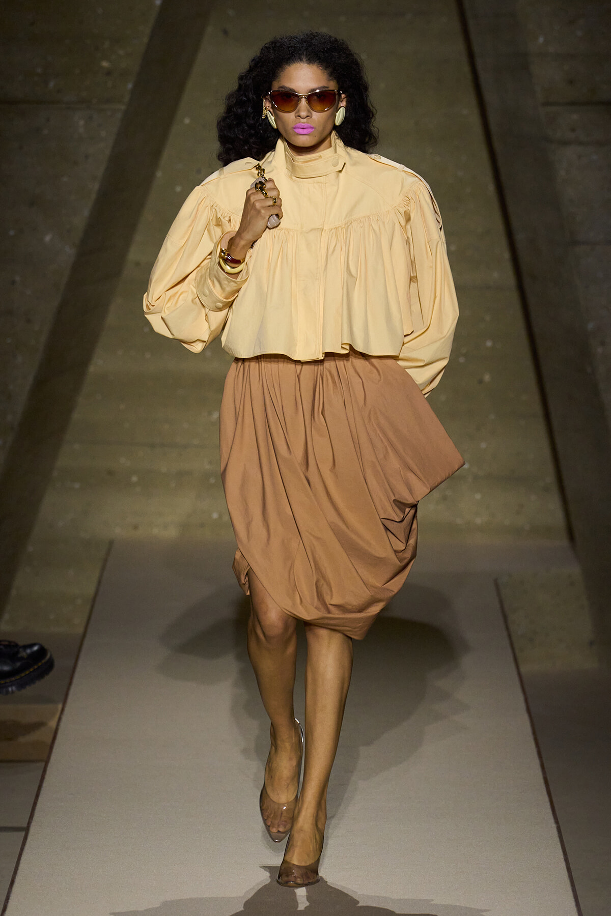 Model walking a runway in a pale yellow, voluminous blouse and tan bubble skirt, accessorized with sunglasses and large earrings.