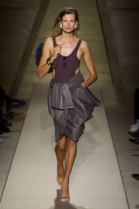 Model walking a runway in a sleeveless plum top and draped gray wrap skirt, pink heels, and white earrings, carrying a blue bag on her shoulder.
