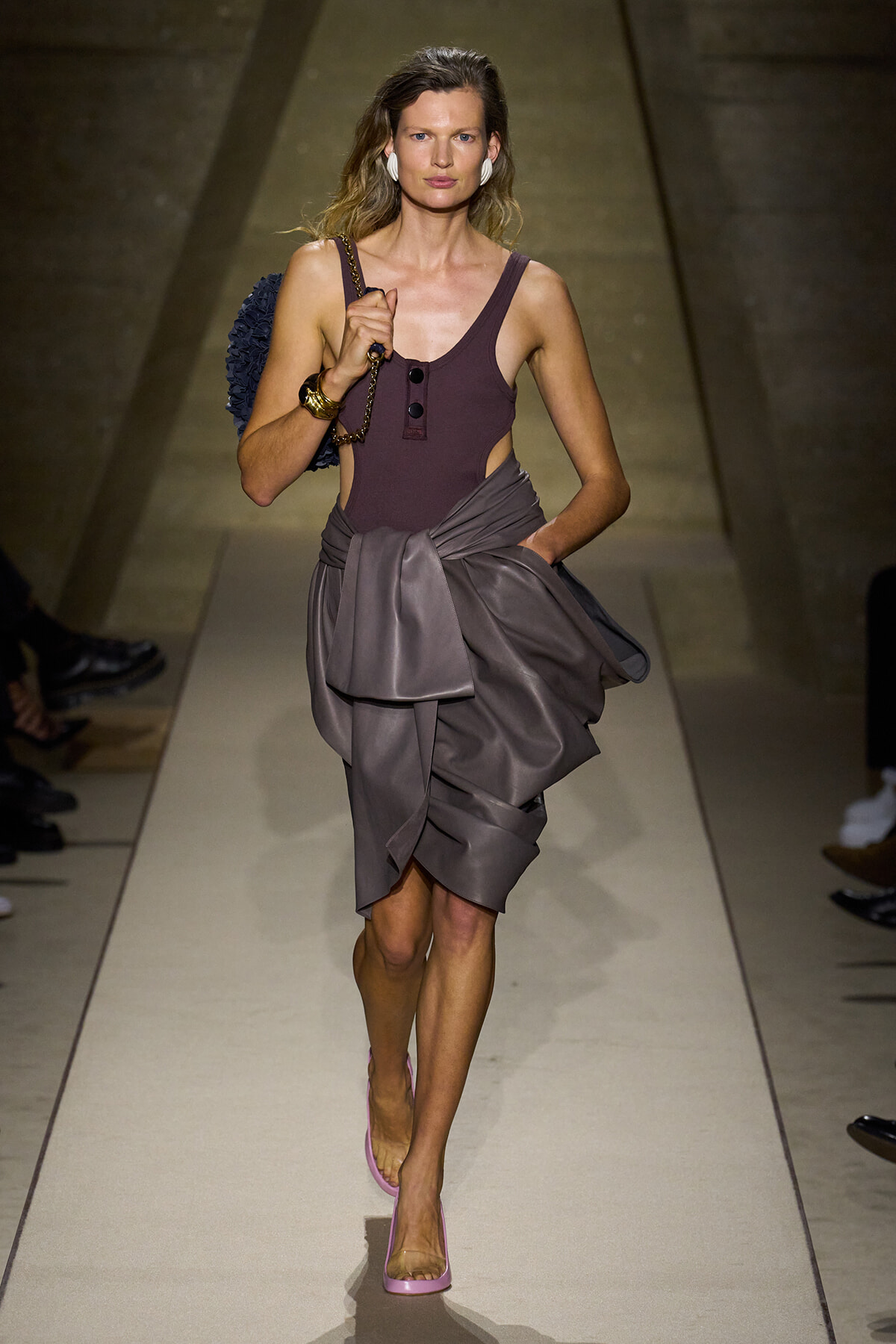 Model walking a runway in a sleeveless plum top and draped gray wrap skirt, pink heels, and white earrings, carrying a blue bag on her shoulder.