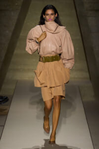 Runway model in a beige coordinated outfit: oversized top with a draped neck, wide olive belt, layered skirt, and clear heels, walking a catwalk.