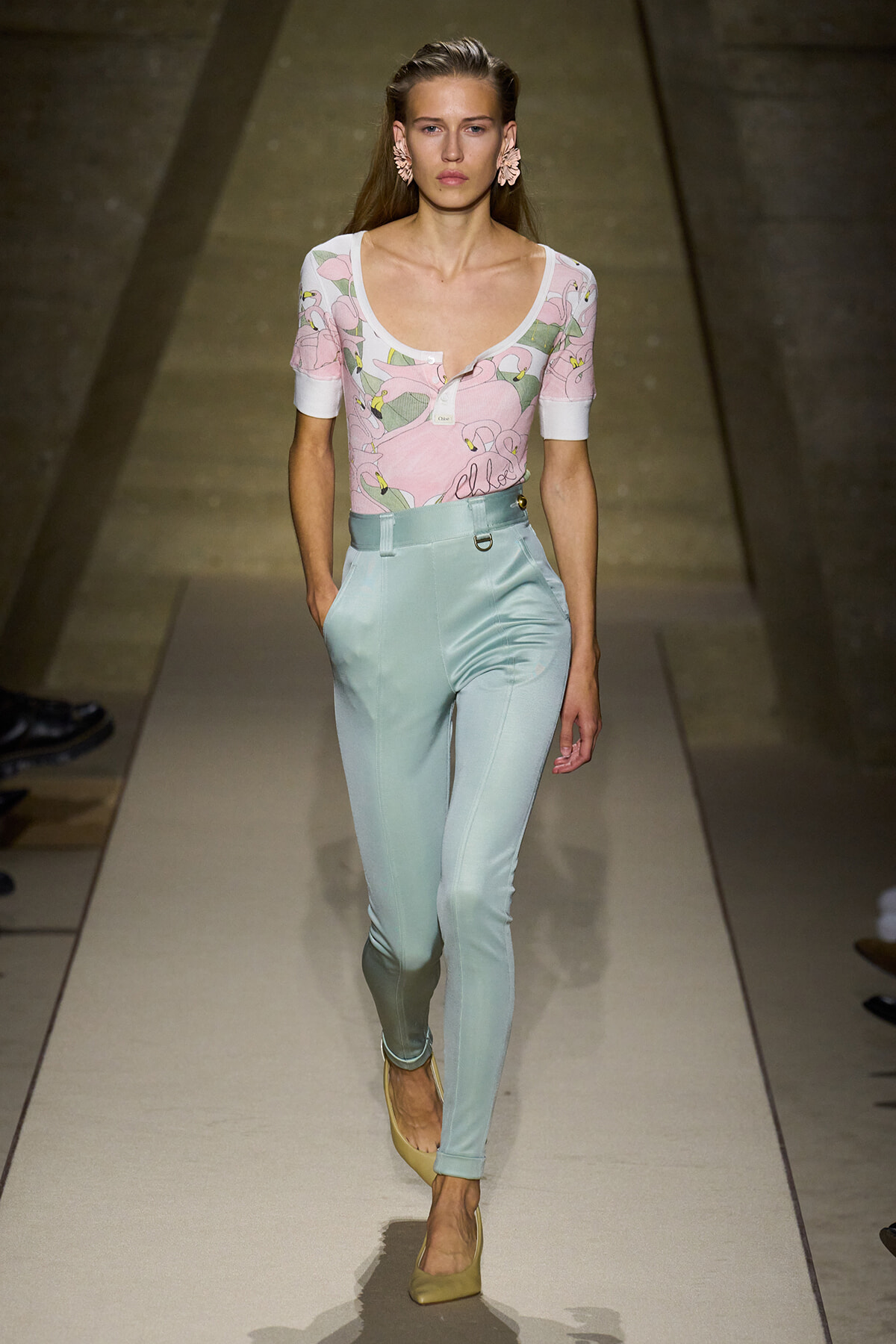 Model walking a runway in a pink floral short-sleeve top and pastel blue high-waisted pants with beige heels.