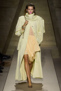 Model walking a runway in a pale yellow trench over a peach dress, holding a gold chain bag.