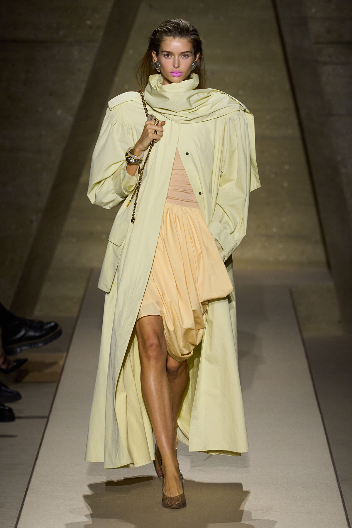 Model walking a runway in a pale yellow trench over a peach dress, holding a gold chain bag.
