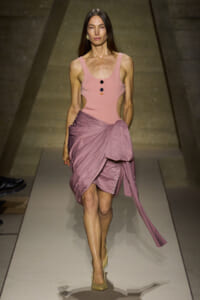 Model walking a fashion runway in a pink sleeveless bodysuit with cutouts and a mauve draped wrap skirt, beige heels.