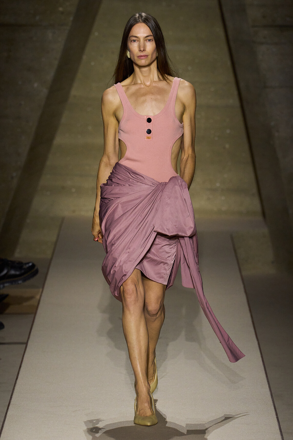 Model walking a fashion runway in a pink sleeveless bodysuit with cutouts and a mauve draped wrap skirt, beige heels.