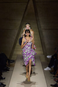 Model walks a beige runway in a purple floral dress with a ruffled skirt, beige heels, and bold earrings, as other models follow in the background.