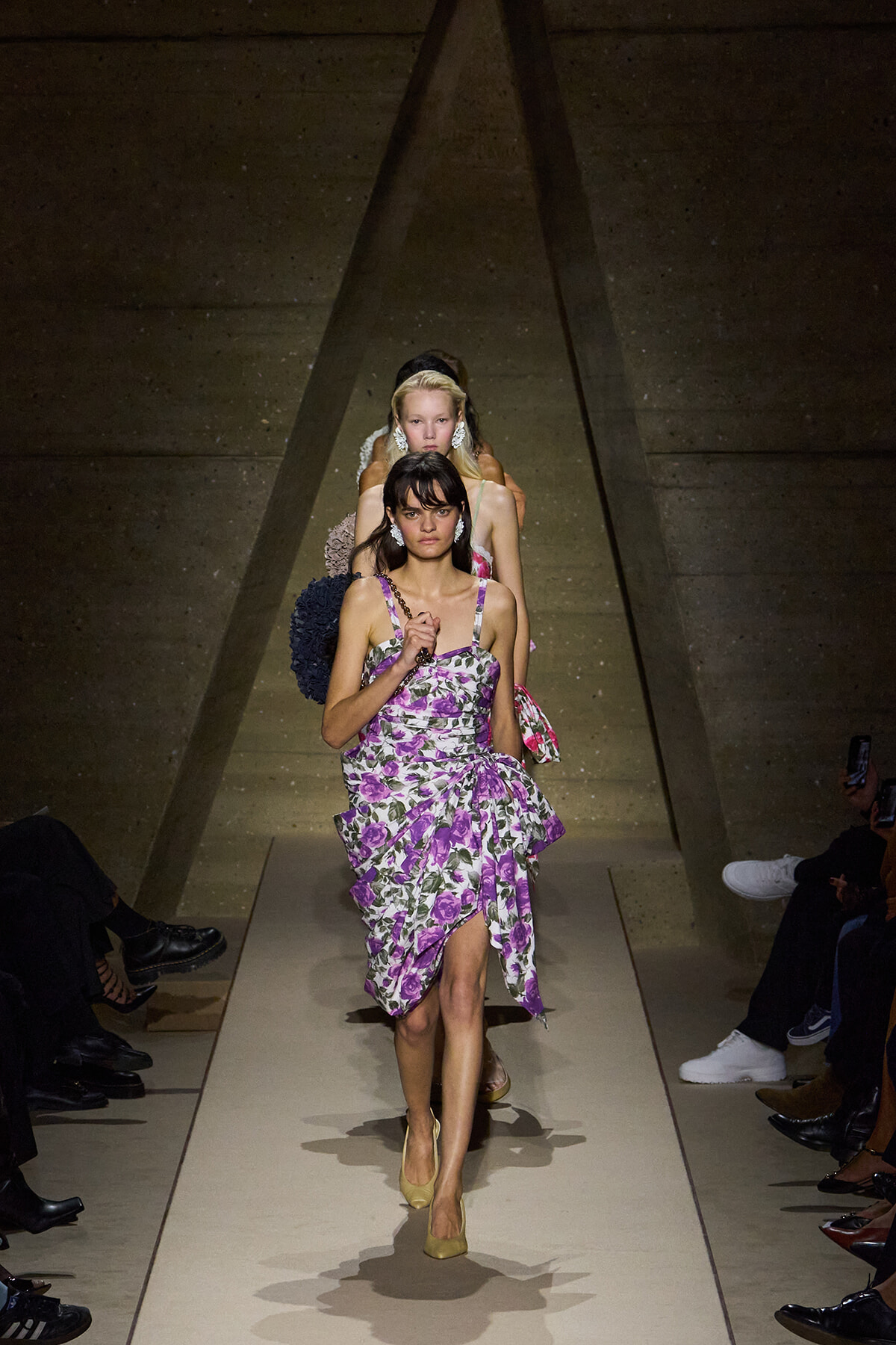Model walks a beige runway in a purple floral dress with a ruffled skirt, beige heels, and bold earrings, as other models follow in the background.