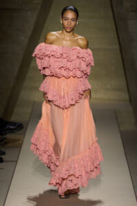 Model walking a runway in a pink off-the-shoulder dress with multiple tiers of ruffled lace and a flowing skirt.