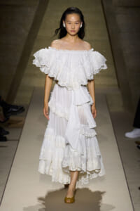 Model walks a runway in a white off-the-shoulder, multi-tiered lace dress with dramatic ruffles and scalloped hems.