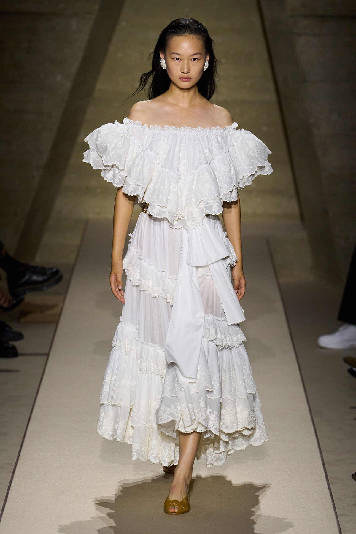 Model walks a runway in a white off-the-shoulder, multi-tiered lace dress with dramatic ruffles and scalloped hems.