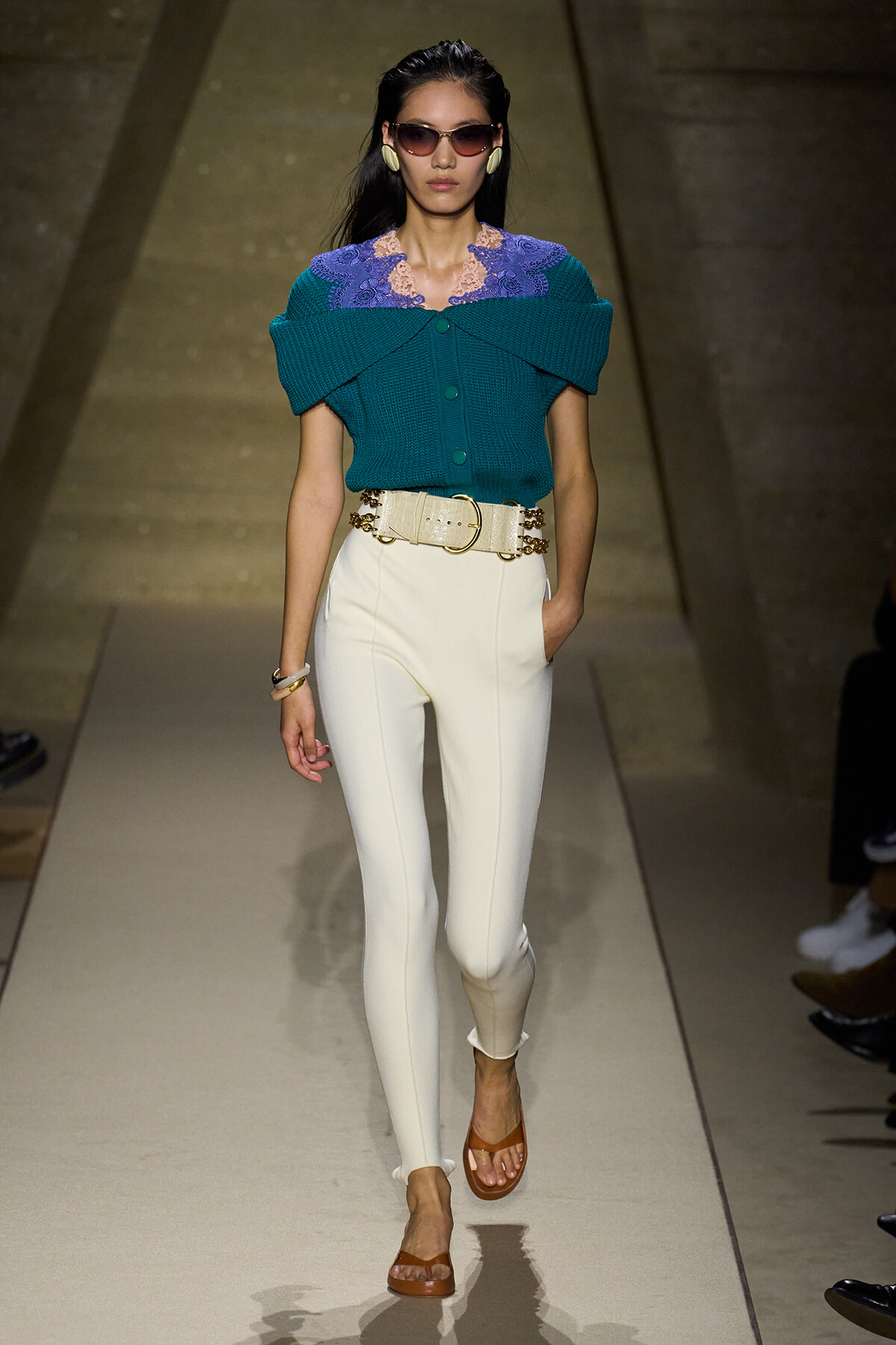 Model walking runway in teal knit top with purple lace collar, white high-waisted pants, and tan sandals.