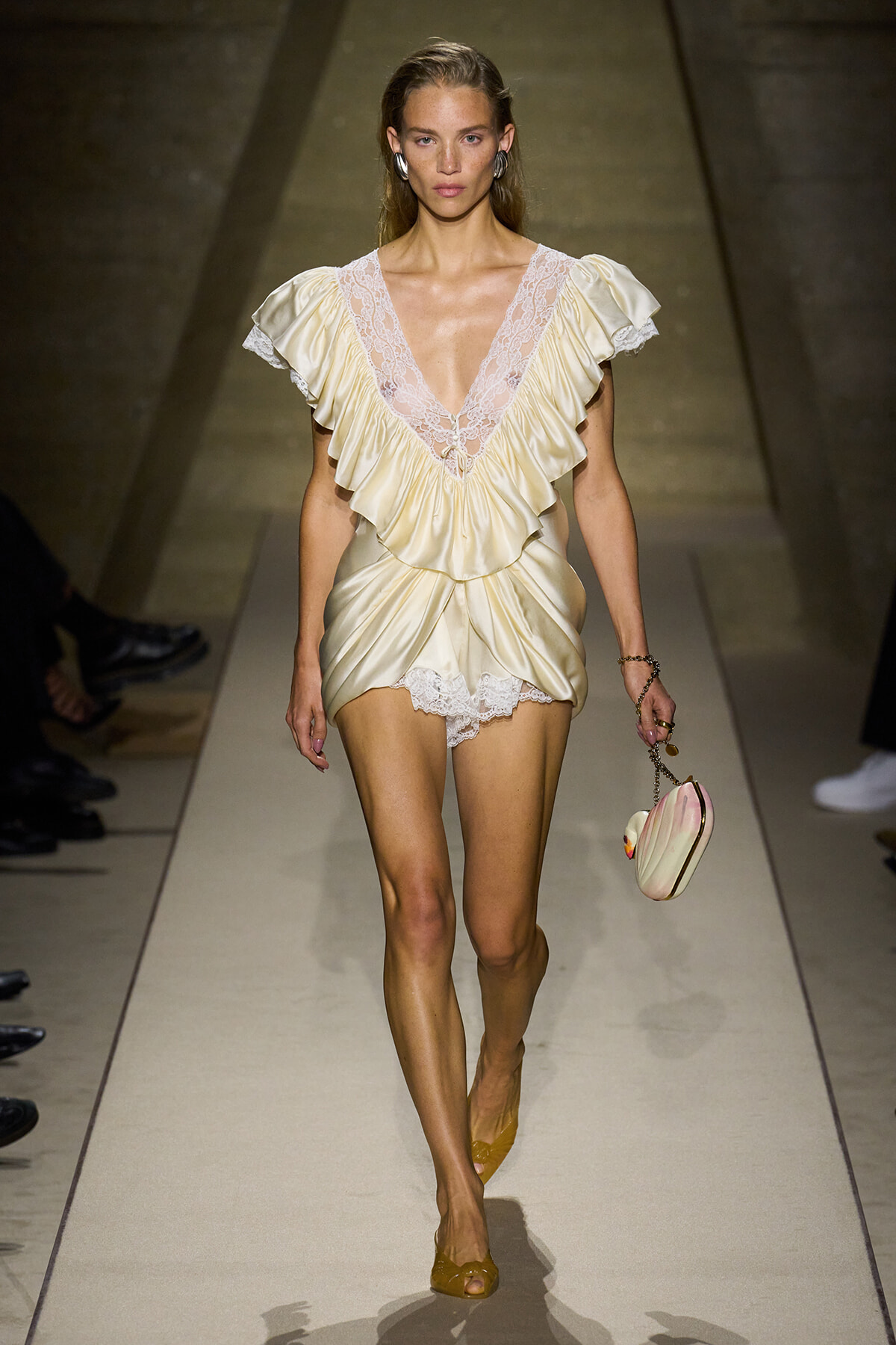 Model walks the runway in a cream satin mini dress with lace V-neck, layered ruffles, and a small round handbag.