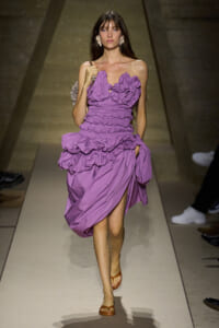 Model walking a runway in a lavender, ruched, ruffled dress with a thigh-high slit and brown sandals, statement earrings visible.