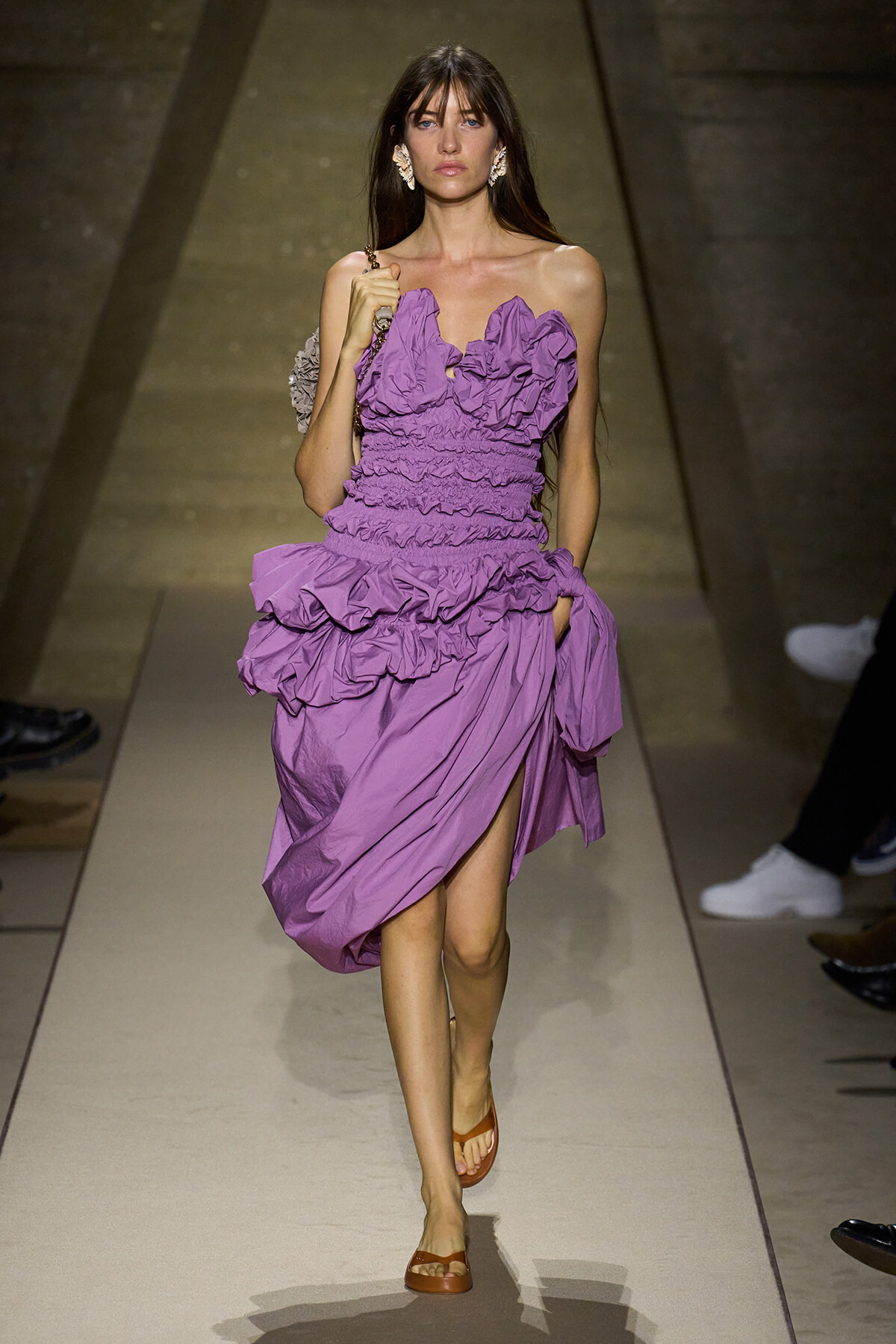Model walking a runway in a lavender, ruched, ruffled dress with a thigh-high slit and brown sandals, statement earrings visible.