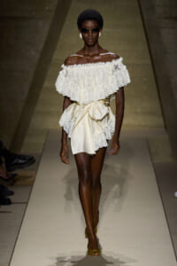 Model walks a runway in an ivory off-the-shoulder lace top with ruffles and a matching satin wrap skirt, sunglasses on.
