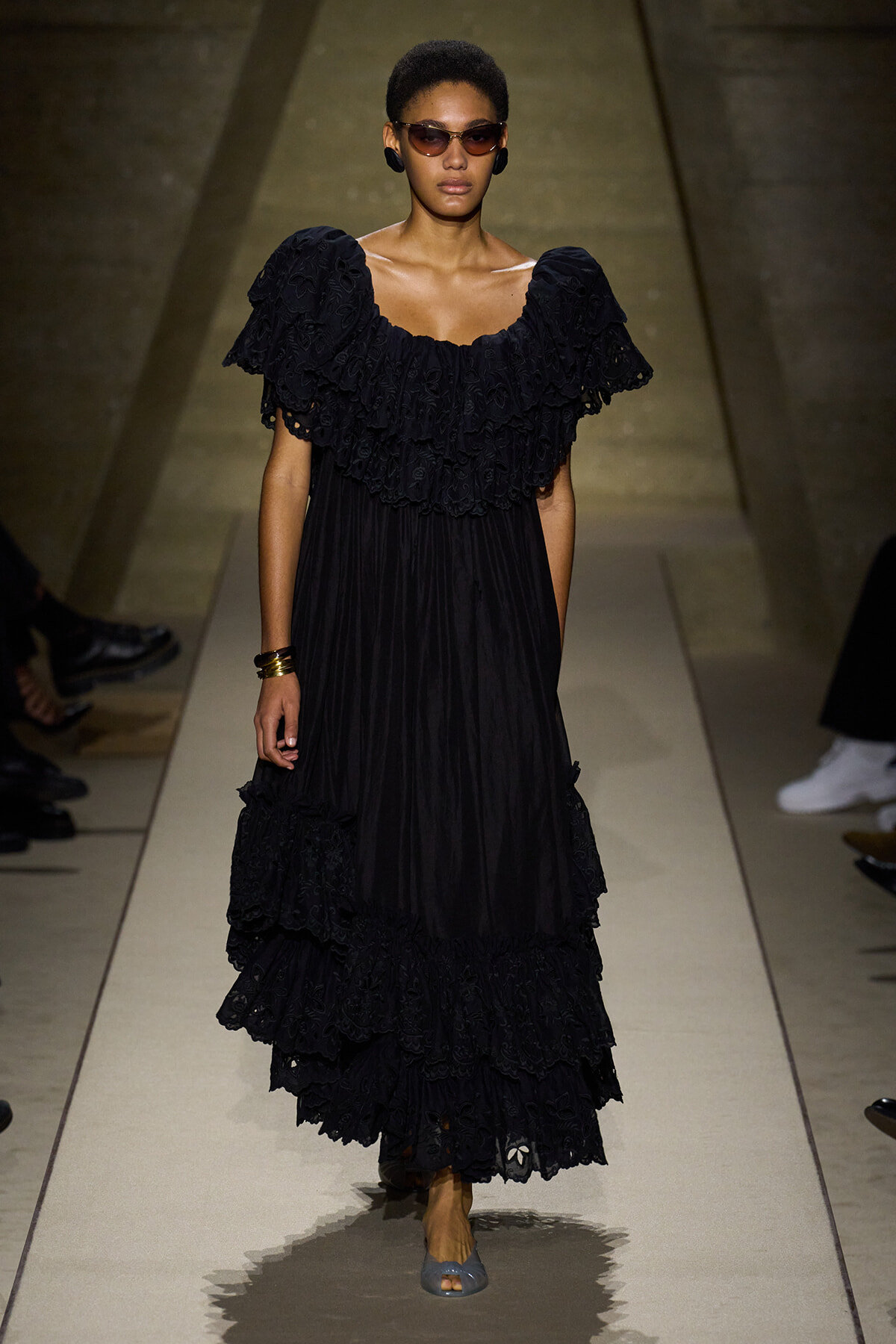 Female model walks a runway in a floor-length black lace dress with voluminous ruffled sleeves and bodice, wearing sunglasses and gold bracelets.