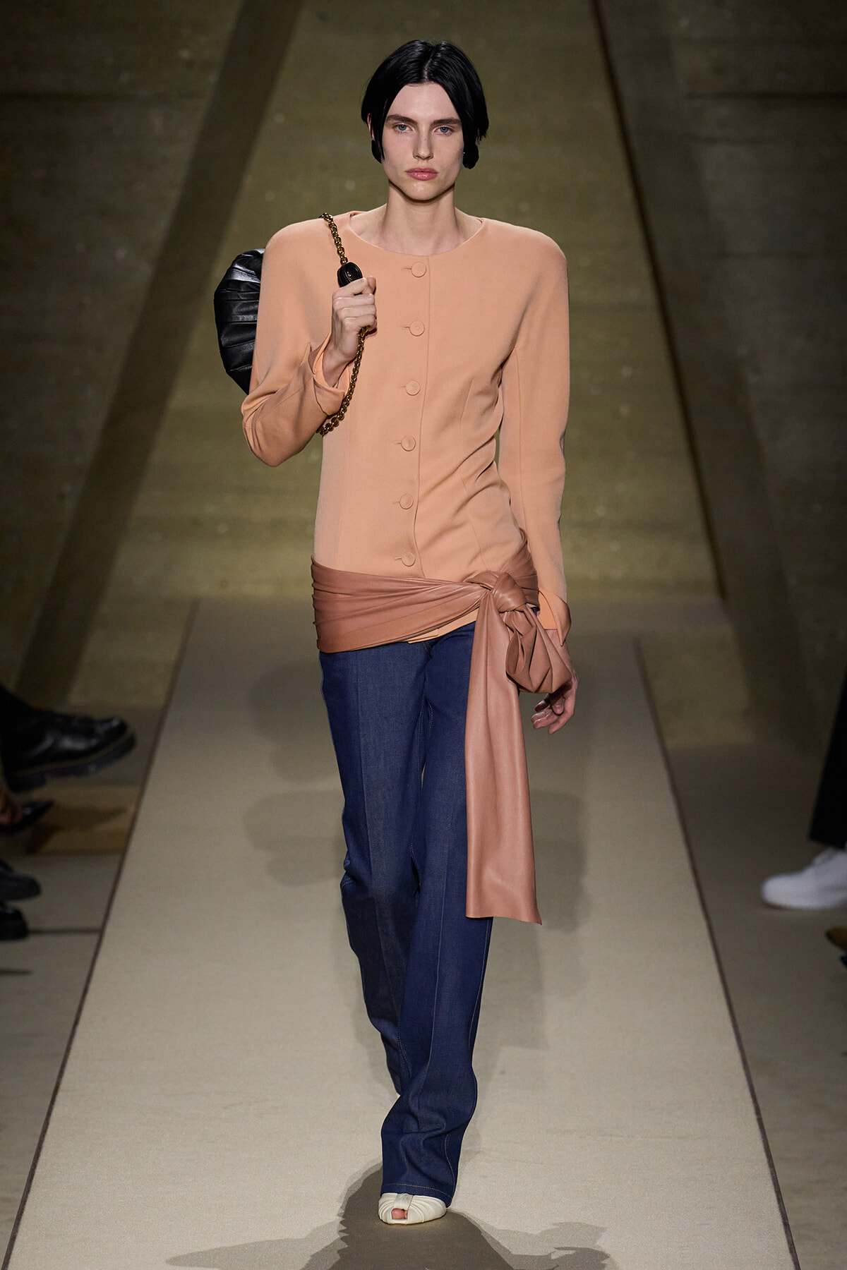 Model walks a fashion runway in a peach long-sleeve buttoned top, wide satin belt tied at the waist, dark blue jeans, and a black shoulder bag.