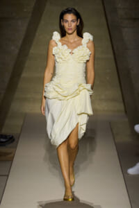 Model walks runway in a pale cream ruffled dress with a structured bodice and draped skirt.