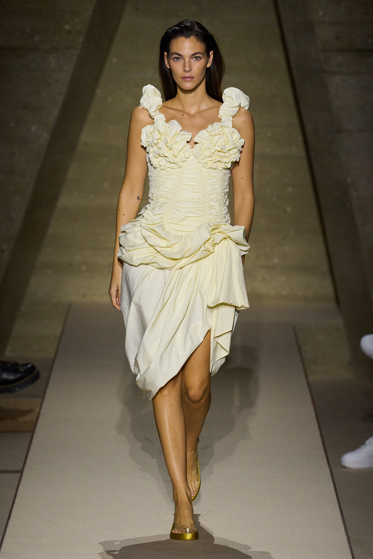 Model walks runway in a pale cream ruffled dress with a structured bodice and draped skirt.