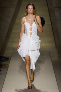 Model walks a fashion runway in a white, knot-front dress with cutouts and a voluminous, draped skirt, carrying a black handbag on her shoulder.