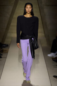Model walking a runway in a black fitted top with structured shoulders and lavender wide-leg pants, cinched with a black leather sash