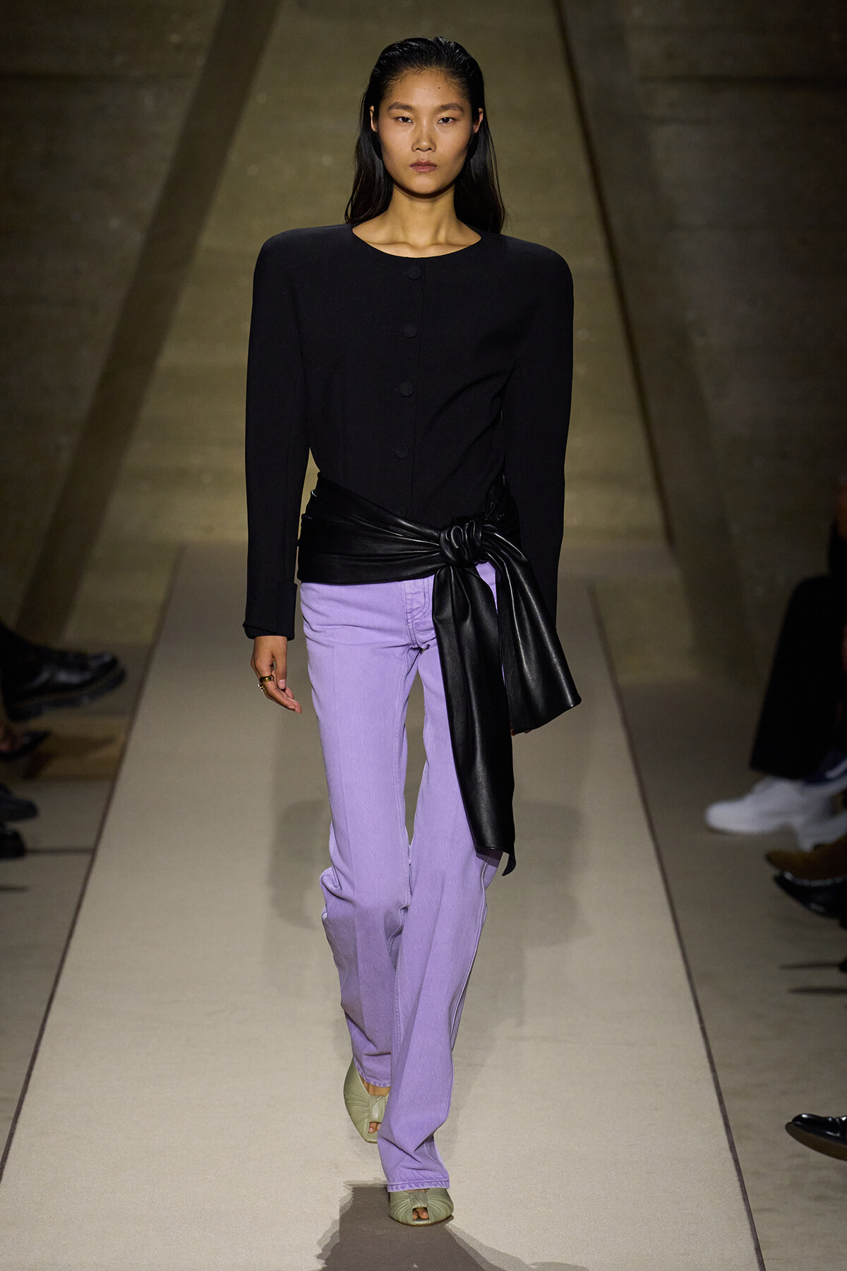 Model walking a runway in a black fitted top with structured shoulders and lavender wide-leg pants, cinched with a black leather sash