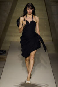 Model walking a runway in a black halter-neck dress with a draped skirt and a chain-strap bag over her shoulder