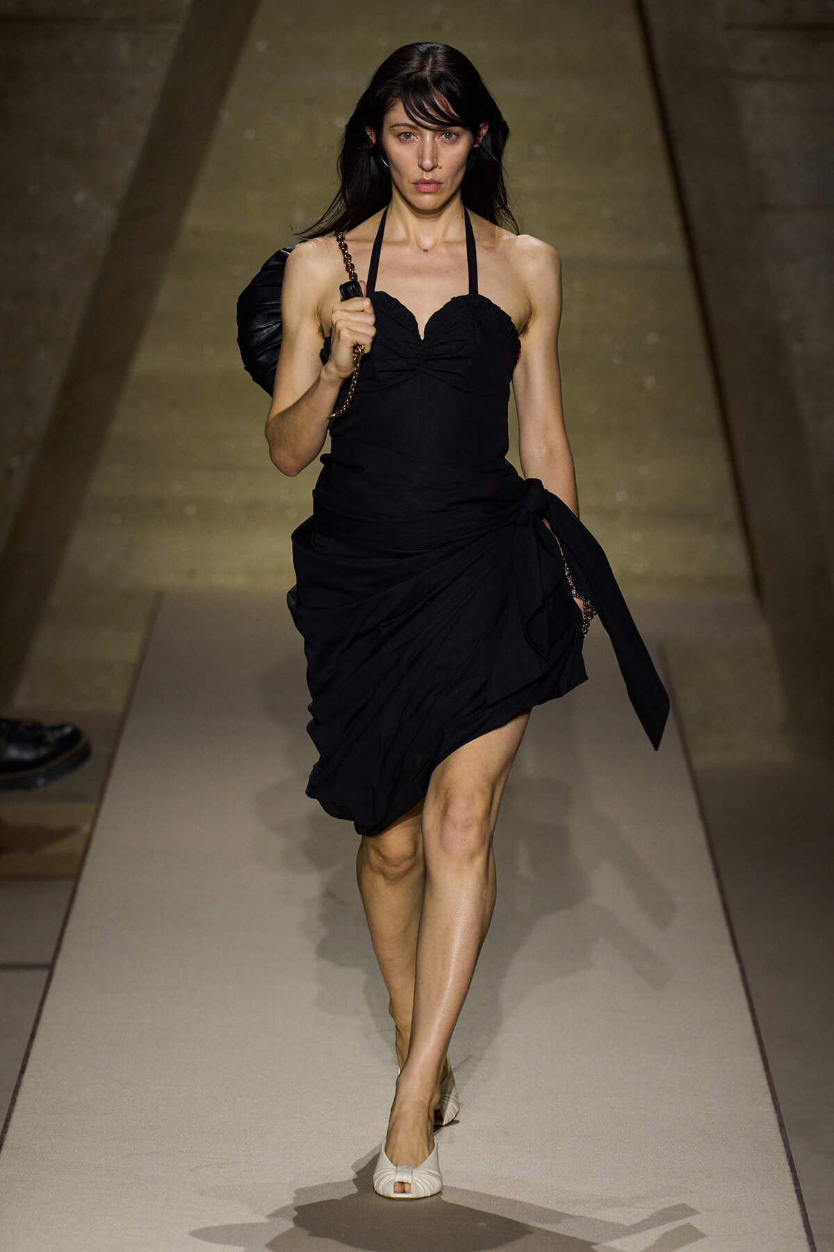 Model walking a runway in a black halter-neck dress with a draped skirt and a chain-strap bag over her shoulder