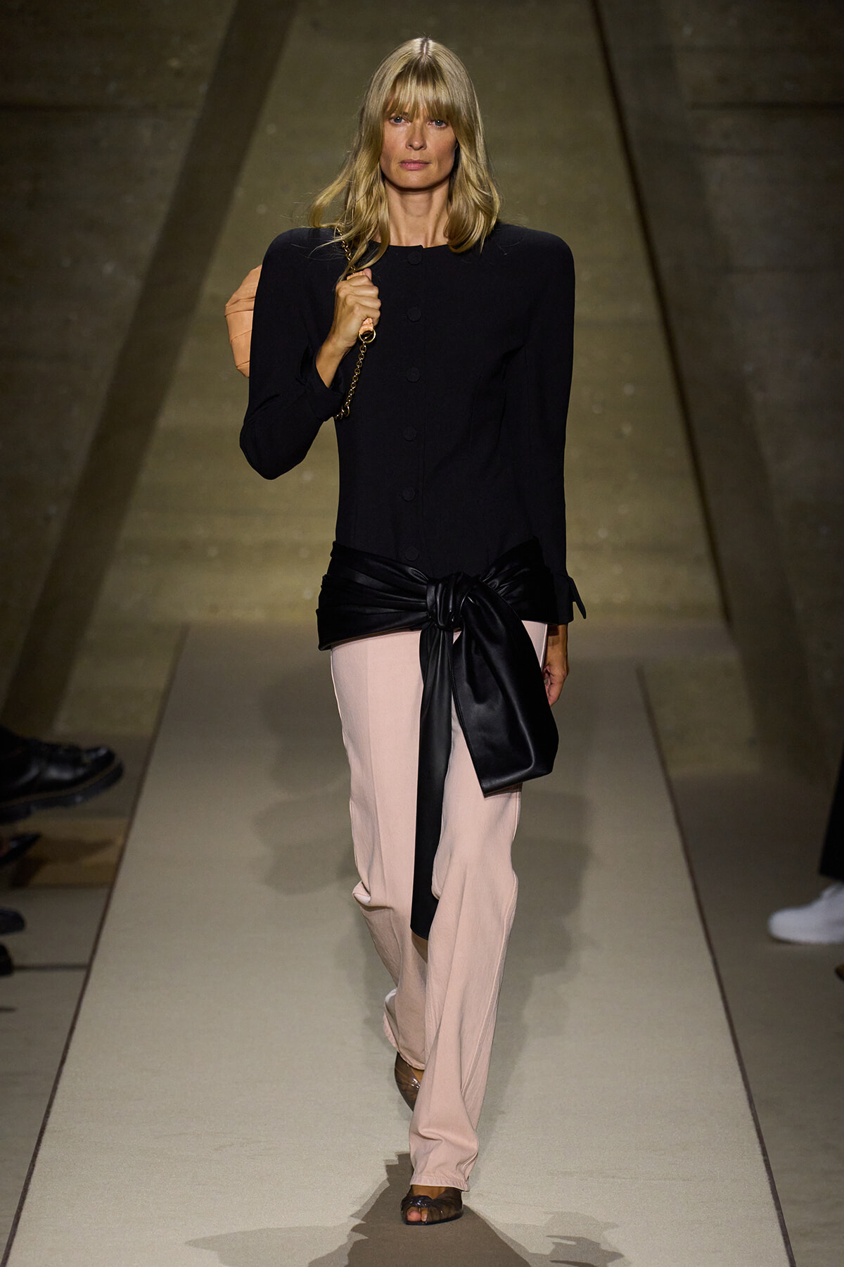 Model walks a fashion runway wearing a black structured top, pale pink wide-leg pants with a black sash, and a quilted handbag over her shoulder.